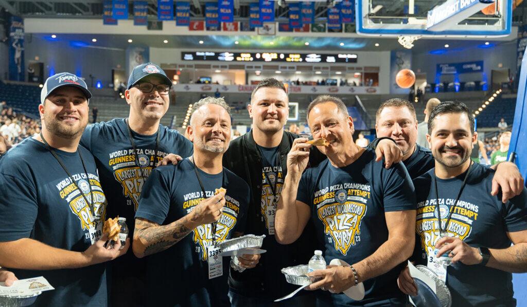 Seven people in matching t-shirts pose for the camera, each has a slice of pizza. The basketball court behind them.