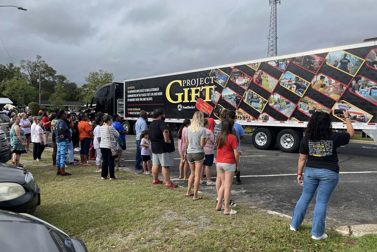 People waiting on a grassy area, looking at a semitruck with "Project Gift" on the side.