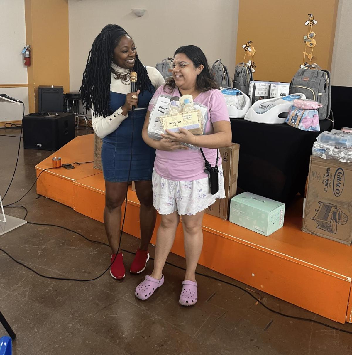 A person with a microphone speaking to another who is holding baby gift items.