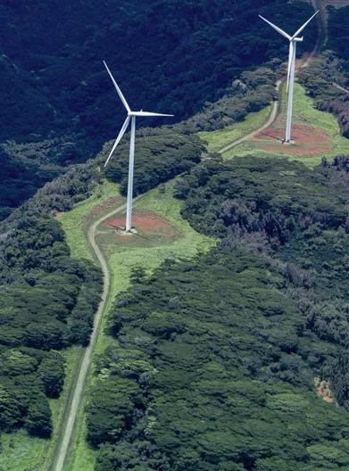 Aerial view of two wind turbines in a mountainous, tree-dense area.