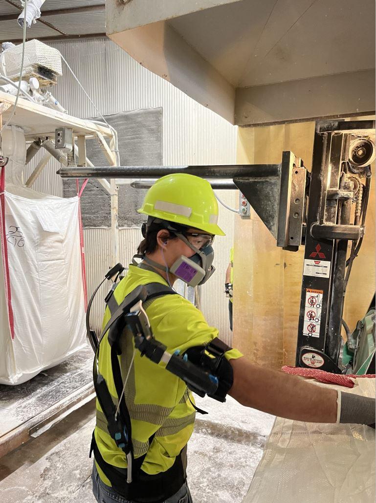 A person in protective gear in a manufacturing area wearing an 'exoskeleton' on their back.