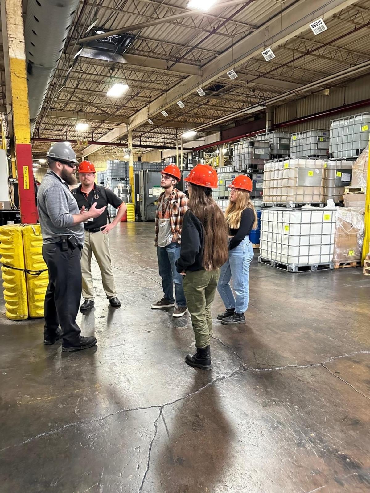 Staff and children in safety hats talking in a warehouse.