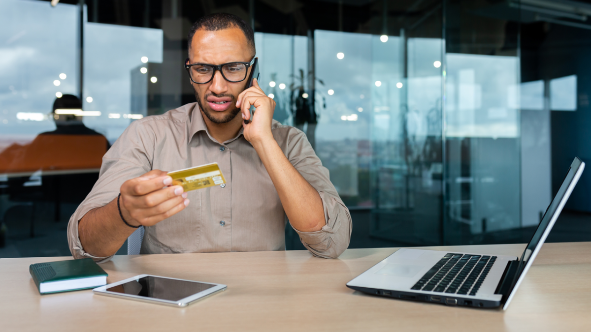 A person talking on a cell phone, looking concerned, holding up a credit card. A laptop computer open next to them on a desk.