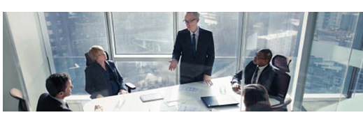 One person standing in a conference room in a tall building, others seated at the table.