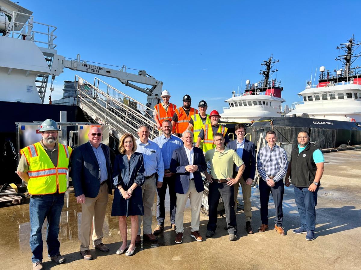 A large group of people standing on a pier in front of a large ship. Some in business attire and some in high-vis and protective wear