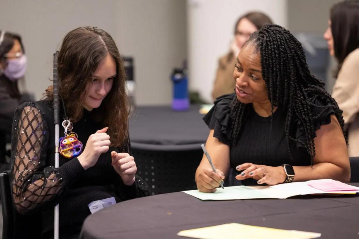 A person talking to another and writing in a folder. The other holds a cane and is smiling.