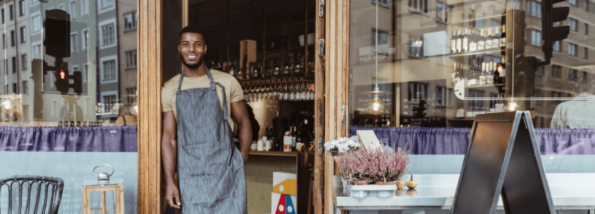 A smiling person wearing an apron, leaning against the door frame of a business.