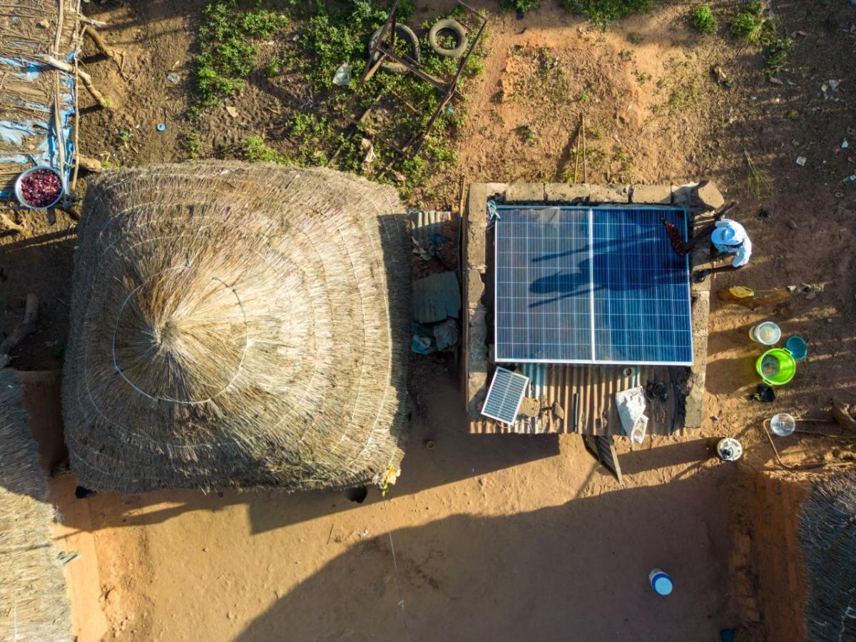 Aerial view of a straw-roof building next to a smaller building with solar panels. A dusty terrain surrounding.