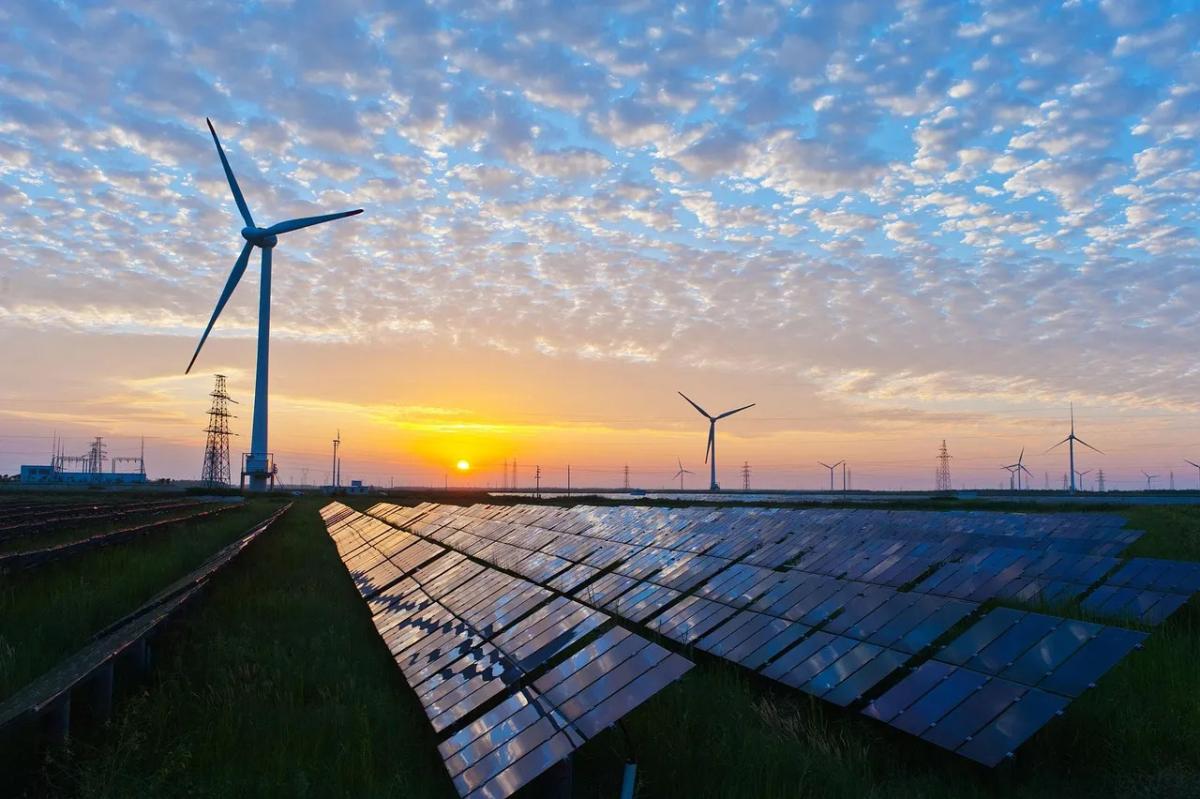 Rows of solar panels with wind turbines and a setting sun in the distance. 