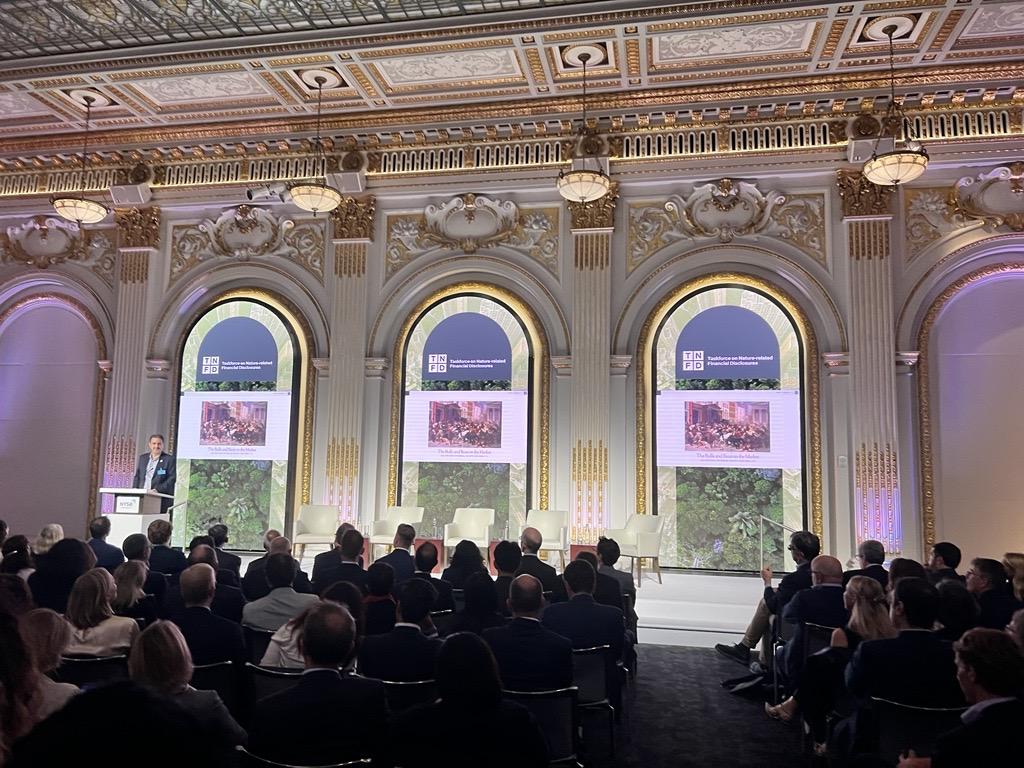 A speaker at a podium in front of a room of seated people. Digital displays behind them,