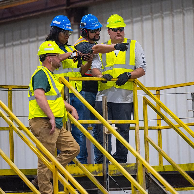 Four employees in hard hats and high-vis vests standing on a platform. One pointing to the distance.