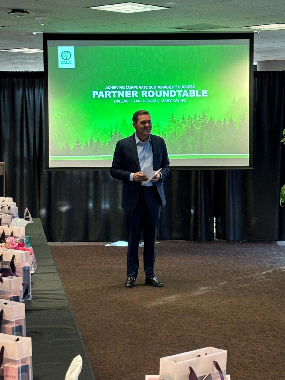 A person standing, speaking to a room of others. A pull-down projector screen "Partner Roundtable" shown.