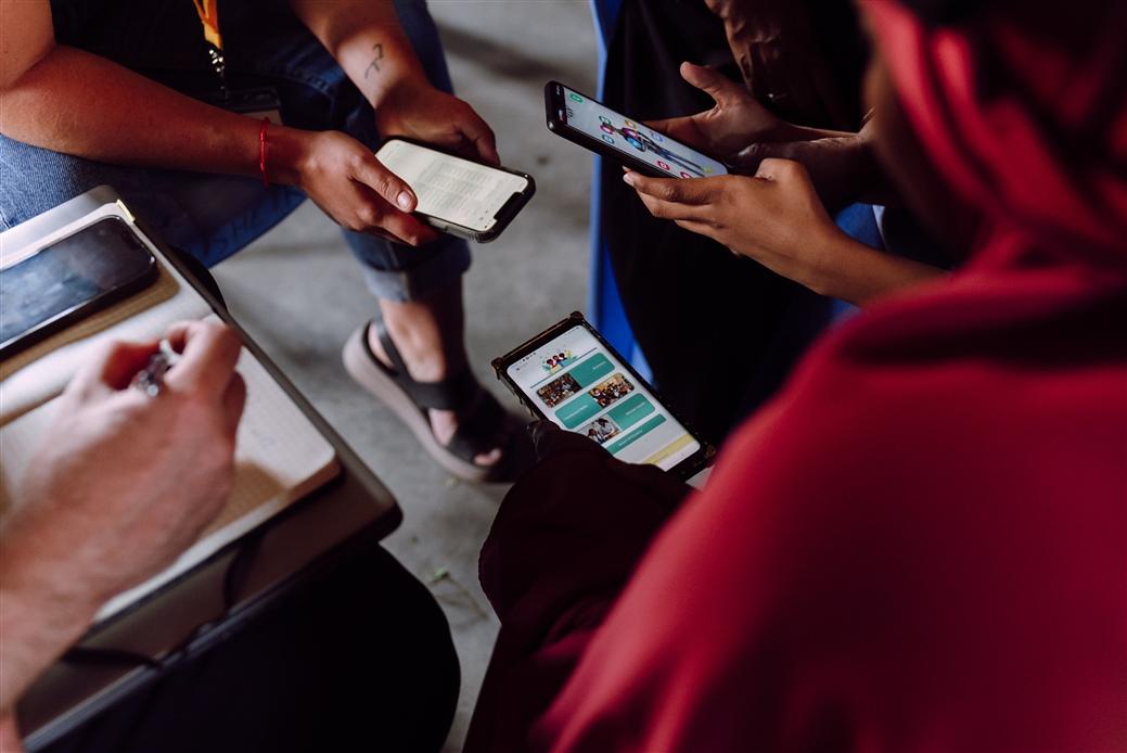 three sets of hands holding electronic devices, a fourth taking notes