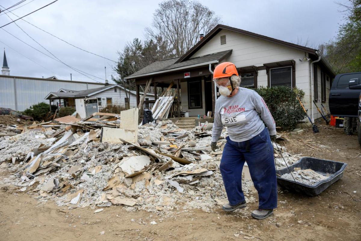 A volunteer in hard hat and respirator pulling a black sled full of rubble. A pile of rubble in front of the house behind them.