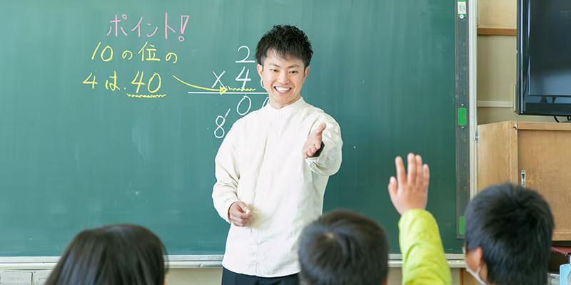 A teacher in front of a chalkboard, pointing to a student.