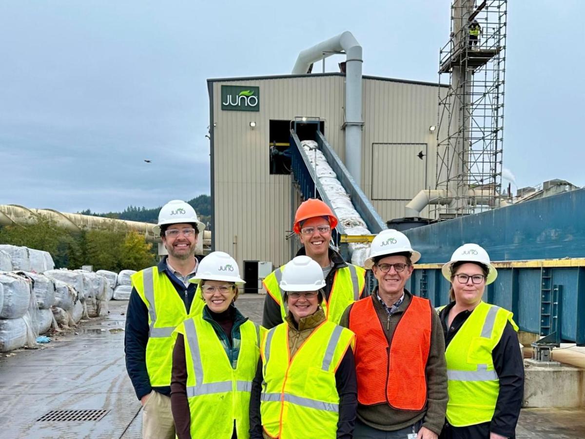 A group of posed people in safety vests and hard hats outside a large facility.