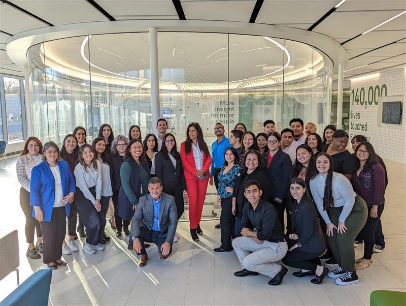 A group of people posed for the camera, some crouching in front, a large glass circular enclosure behind them in an office setting.