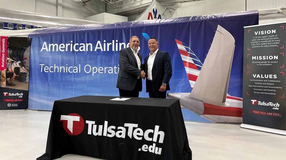 Two people shaking hands in front of an American Airlines banner, behind a Tulsa Tech stand 