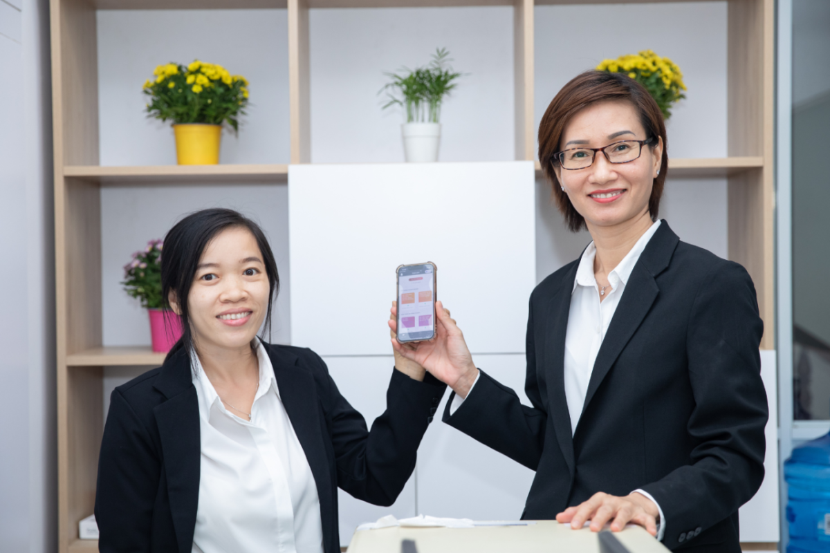 Two people holding the same cell phone. A bookcase behind them with a plant in each cube.