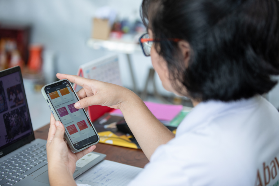 Looking over a person's shoulder, they are using the HerVenture app on a cell phone. A laptop open in front of them on a desk.