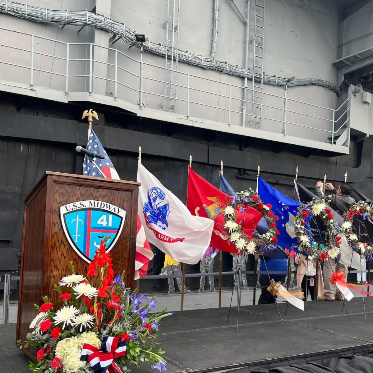 A Stage and empty podium, many flags and a large ship behind it. A flower arrangement in front of the podium.