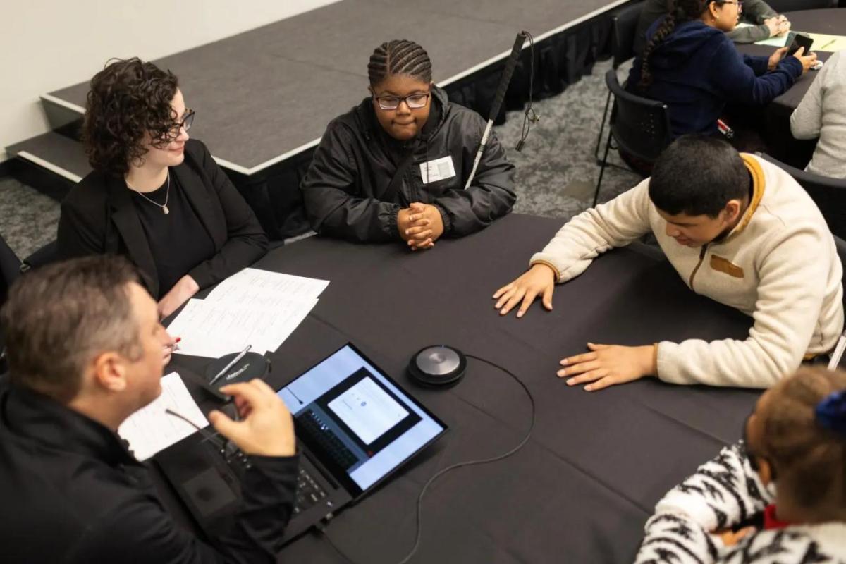 A table with five people seated. One with an open laptop pointed to the others.