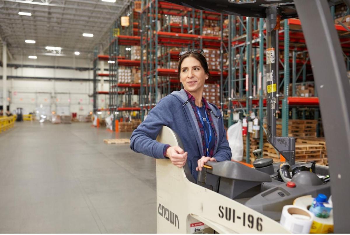 A woman in a warehouse stands in the operators seat of a forklift. She looks over her right shoulder and smiles with floor to ceiling shelves stacked with boxes behind her.