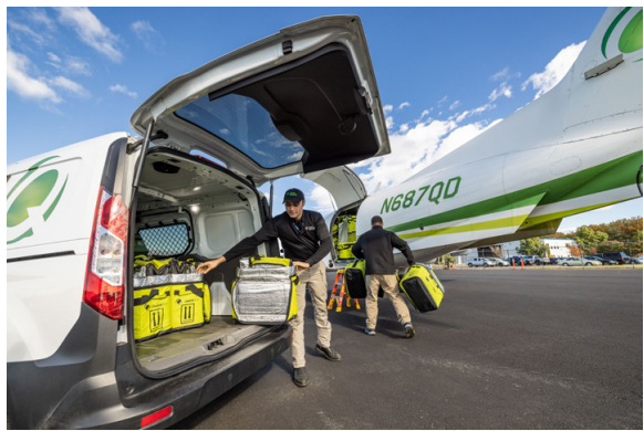 Two people unloading boxes from the back of a van, headed towards a small plane.