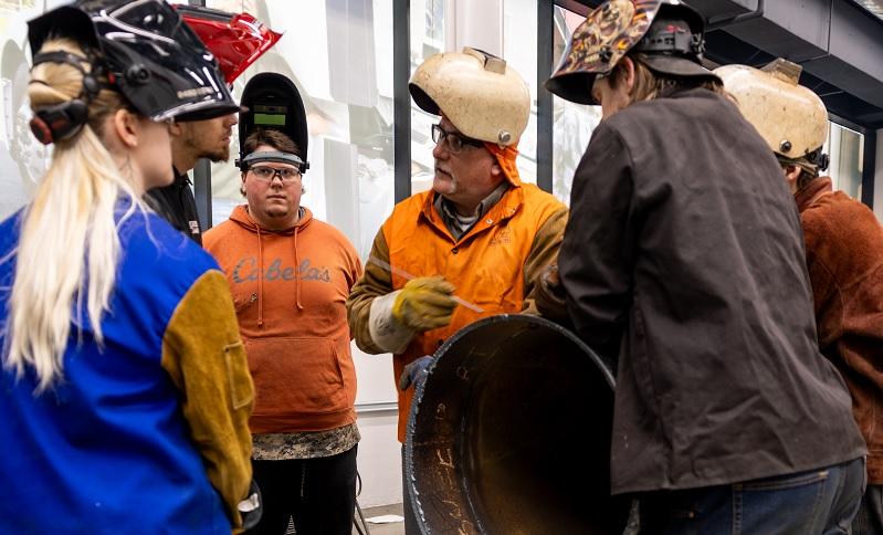 A teacher standing in a circle with students. All wearing welding helmets and protective clothing.