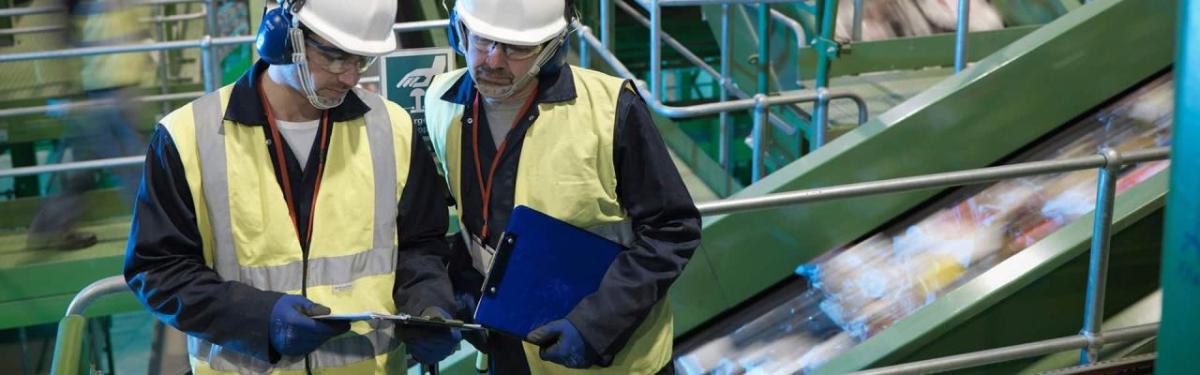 Two people in hard hats, protective wear and high-vis vests looking at a clip board. Fast moving items on a narrow ramp behind them.