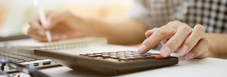 Close up of a person using a calculator, writing with the other hand and a laptop in front of them.