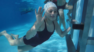 A child underwater waving to the camera. Other children swimming around them.