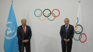 Two people stood in front of an Olympics logo backdrop, talking into a standing microphone 